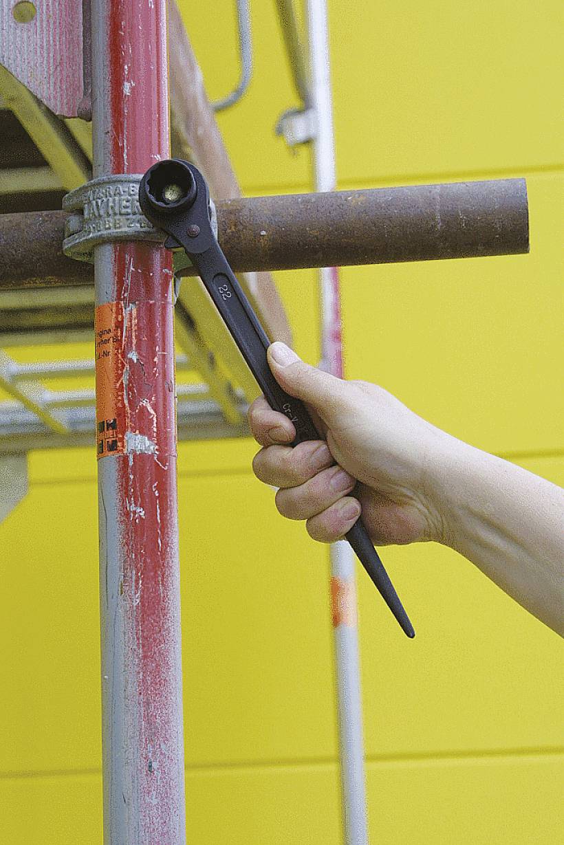 Eine Hand hält einen Schraubenschlüssel und zieht eine Metallstange an einem roten Baugerüst an einer gelben Wand fest.