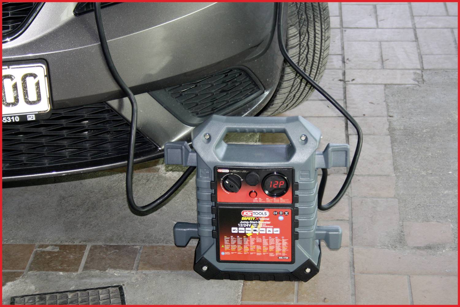 Tragbares Starthilfegerät auf dem Boden vor einem Auto, mit angeschlossenen Kabeln. Display zeigt Batteriespannung von 12,9V an.