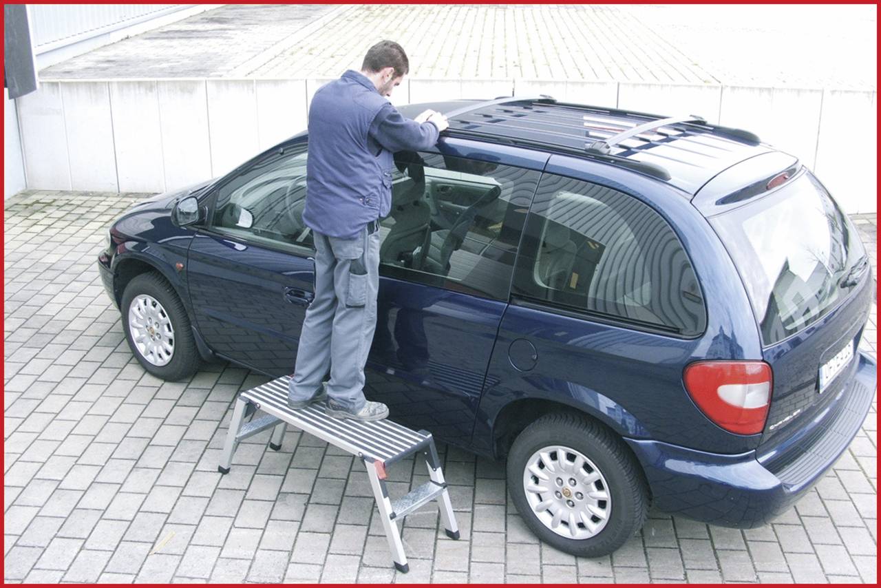 Ein Mann steht auf einer Leiter neben einem blauen Auto und arbeitet am Dach, auf einem gepflasterten Boden vor einem Gebäude.