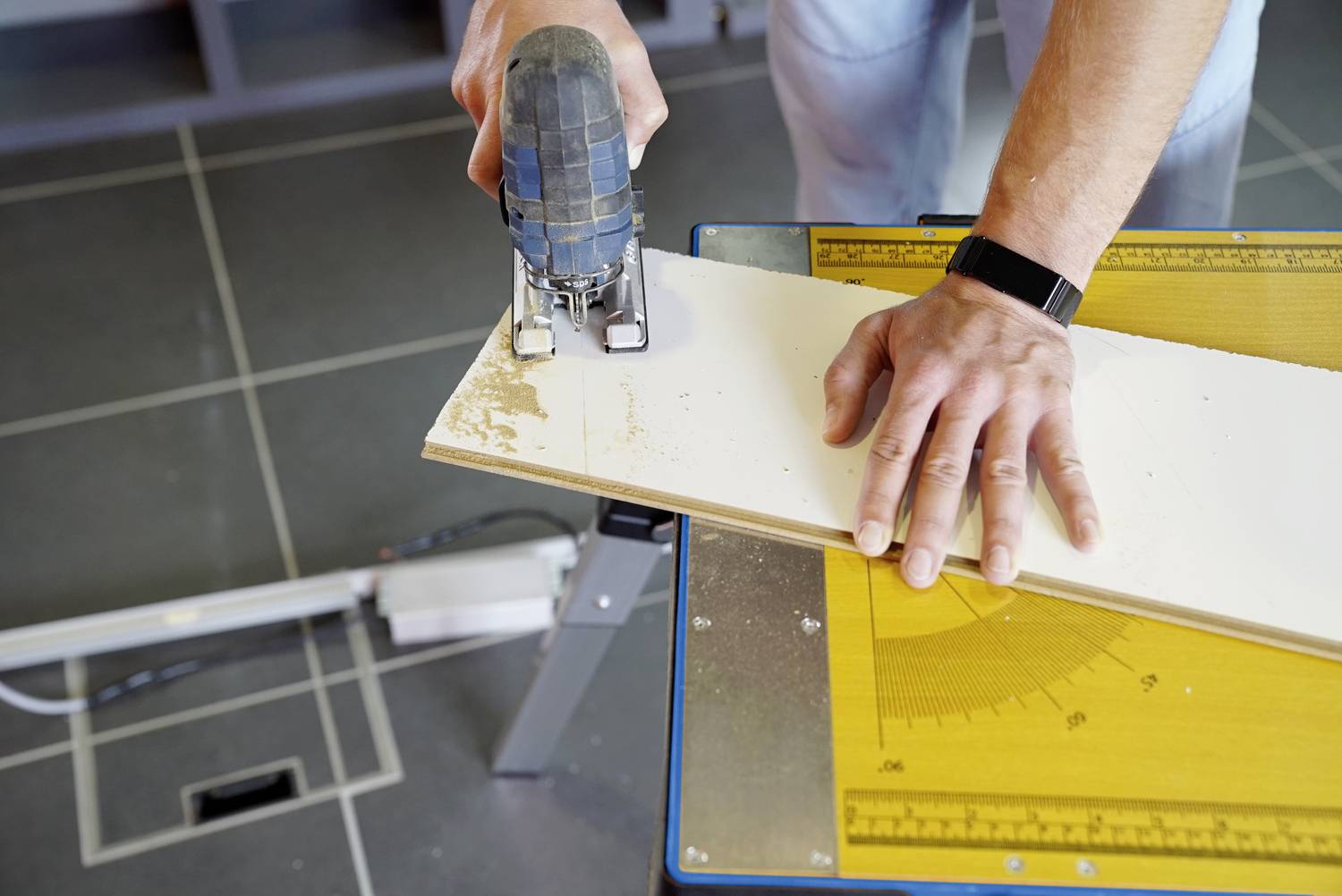 Eine Person schneidet mit einer Stichsäge ein Holzbrett auf einem Tisch mit Messskala. Die Hände halten das Brett fest.