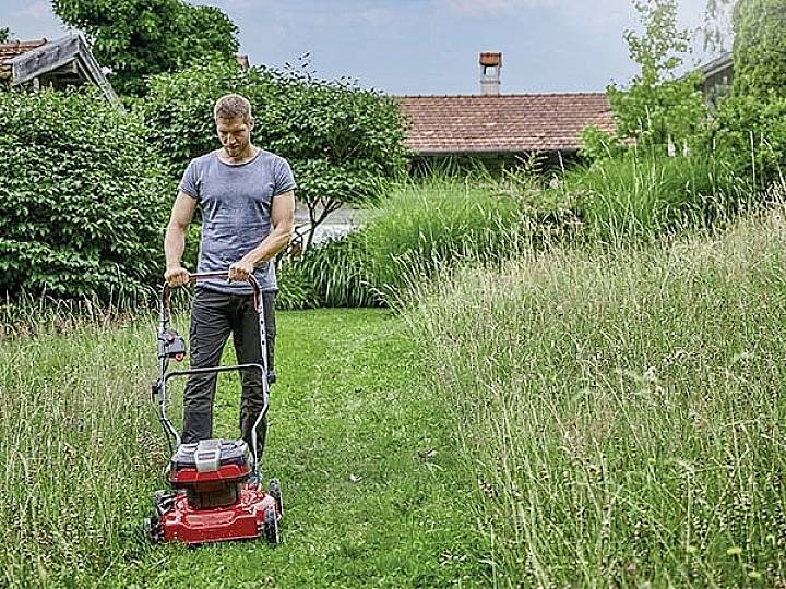 Eine Person mäht mit einem roten Rasenmäher in einem Garten den Rasen. Im Hintergrund sind Bäume und ein Haus zu sehen.