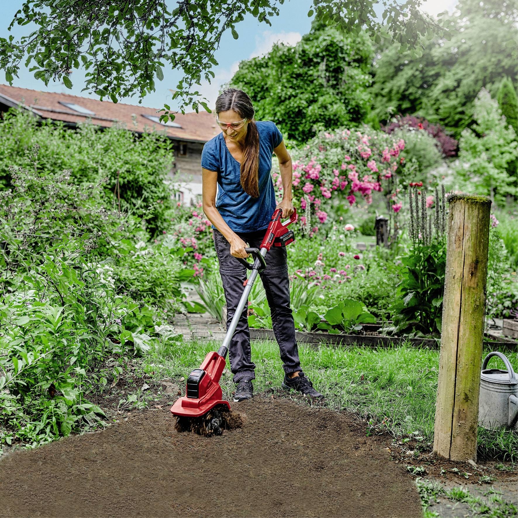 Eine Frau arbeitet im Garten mit einem elektrischen Kultivator. Im Hintergrund sind Blumenbeete und Bäume zu sehen.