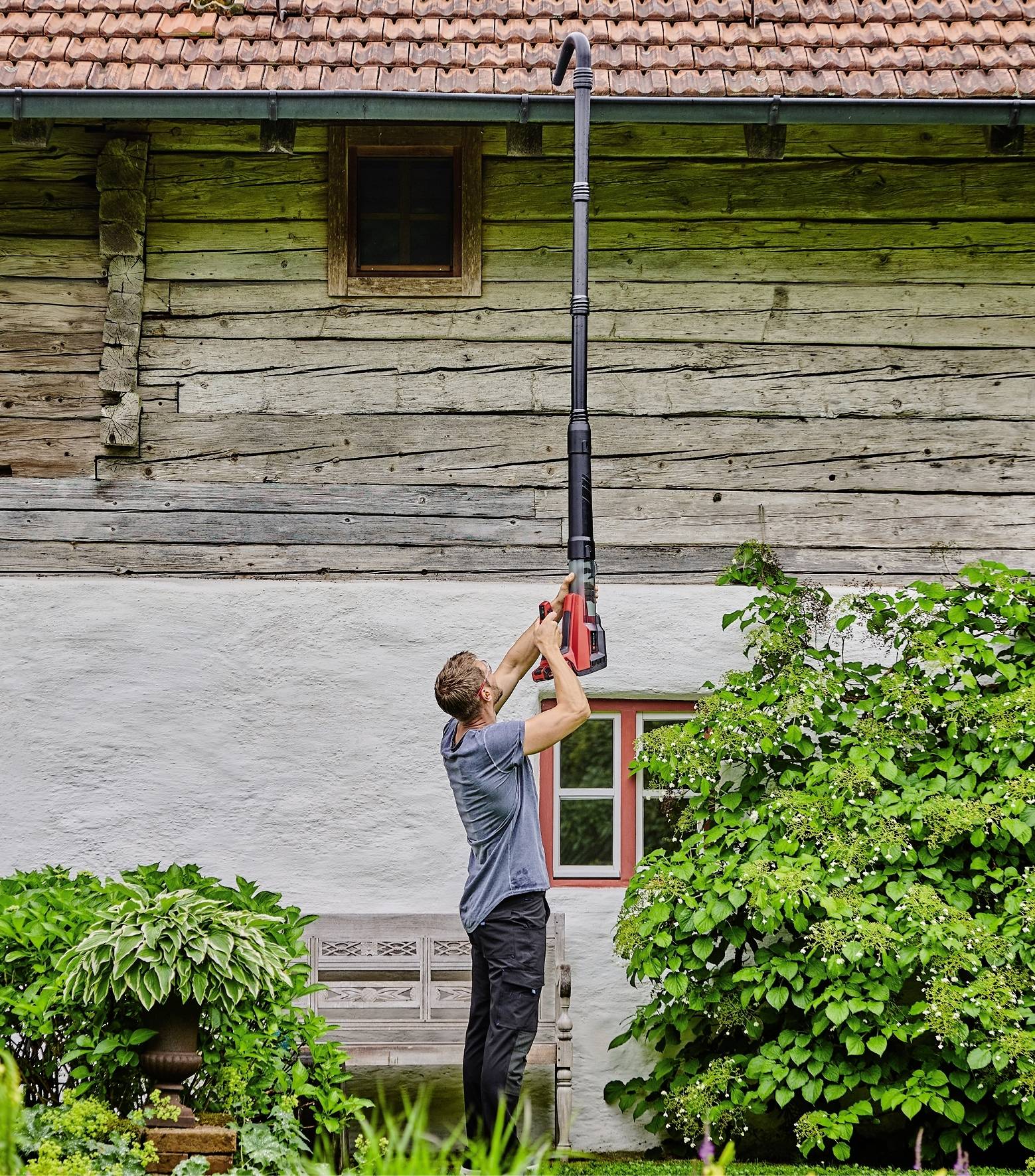 Ein Mann benutzt einen langen Laubbläser, um das Dach eines alten Holzhauses zu reinigen. Im Vordergrund befinden sich grüne Büsche und Pflanzen.