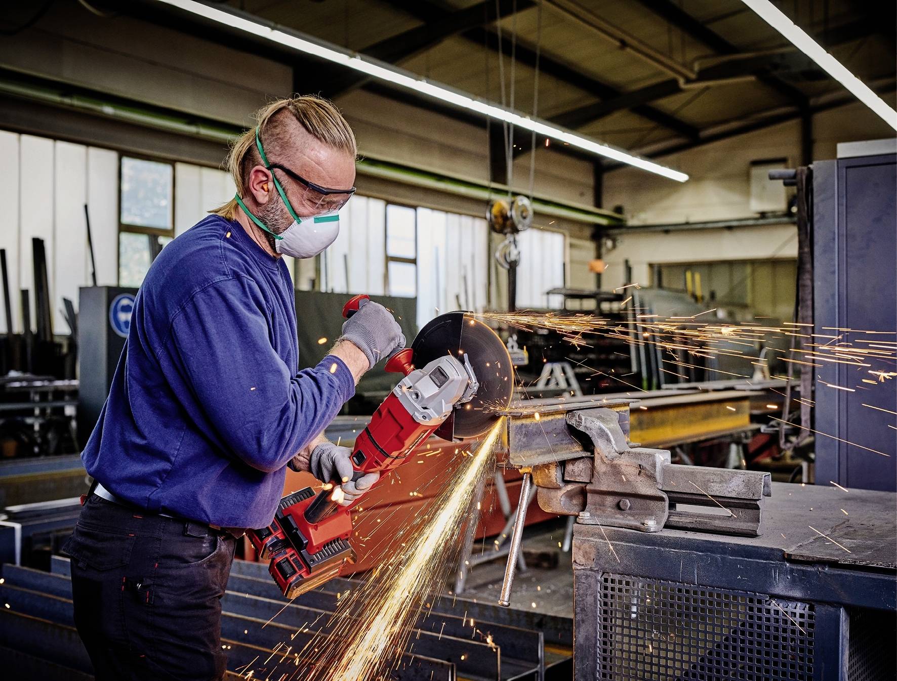Ein Mann mit Schutzbrille und Maske schleift Metall in einer Werkstatt. Funken fliegen vom Schleifgerät, Umgebung industriell.