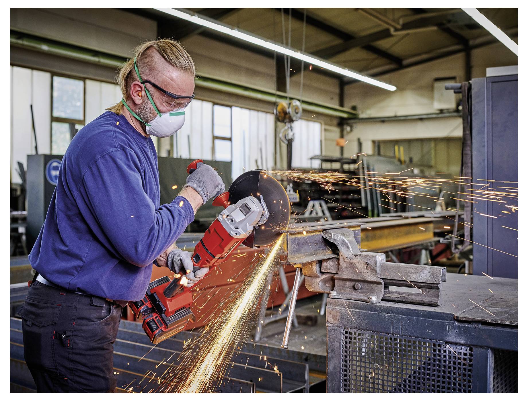 Arbeiter in Schutzausrüstung nutzt einen roten Winkelschleiffer auf Metall in einer Werkstatt und sendet Funken über eine Werkbank.