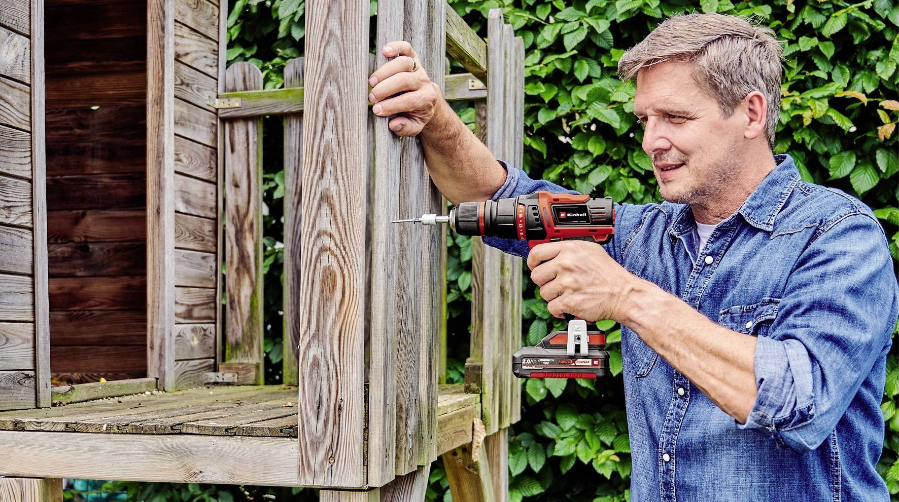 Ein Mann bohrt mit einer elektrischen Bohrmaschine Holzlatten zusammen. Im Hintergrund ist eine Holzkonstruktion zu sehen.