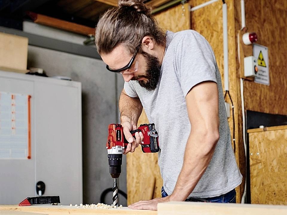 Ein Mann mit Schutzbrille arbeitet mit einem Akkuschrauber an einem Holzbrett in einer Werkstattumgebung.