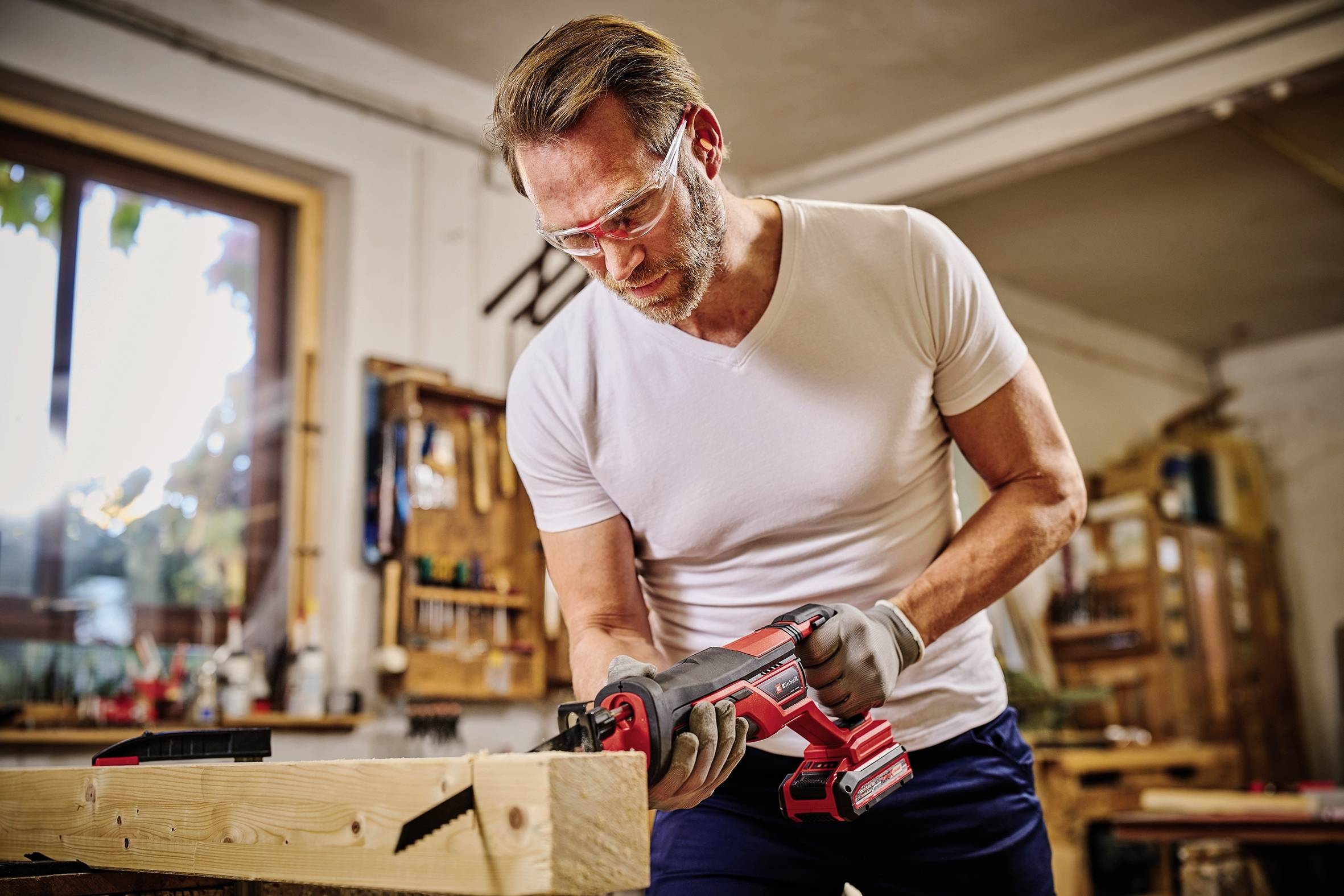 Ein Mann in einer Werkstatt schneidet mit einer Elektrosäge Holz. Er trägt Schutzbrille und Handschuhe und arbeitet konzentriert an einem Holzbalken.