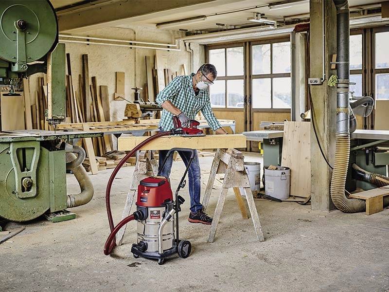 Eine Person arbeitet in einer Tischlerei mit einem Elektrohobel, um ein Holzbrett zu glätten. Im Hintergrund sind Maschinen und Holzvorräte zu sehen.