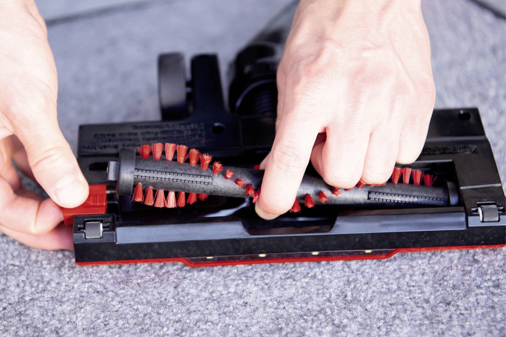 Person, die die Bürstenrolle eines Staubsaugers wechselt. Hände sind sichtbar, wie sie die Bürste aus der Basis herausziehen. Teppich im Hintergrund.