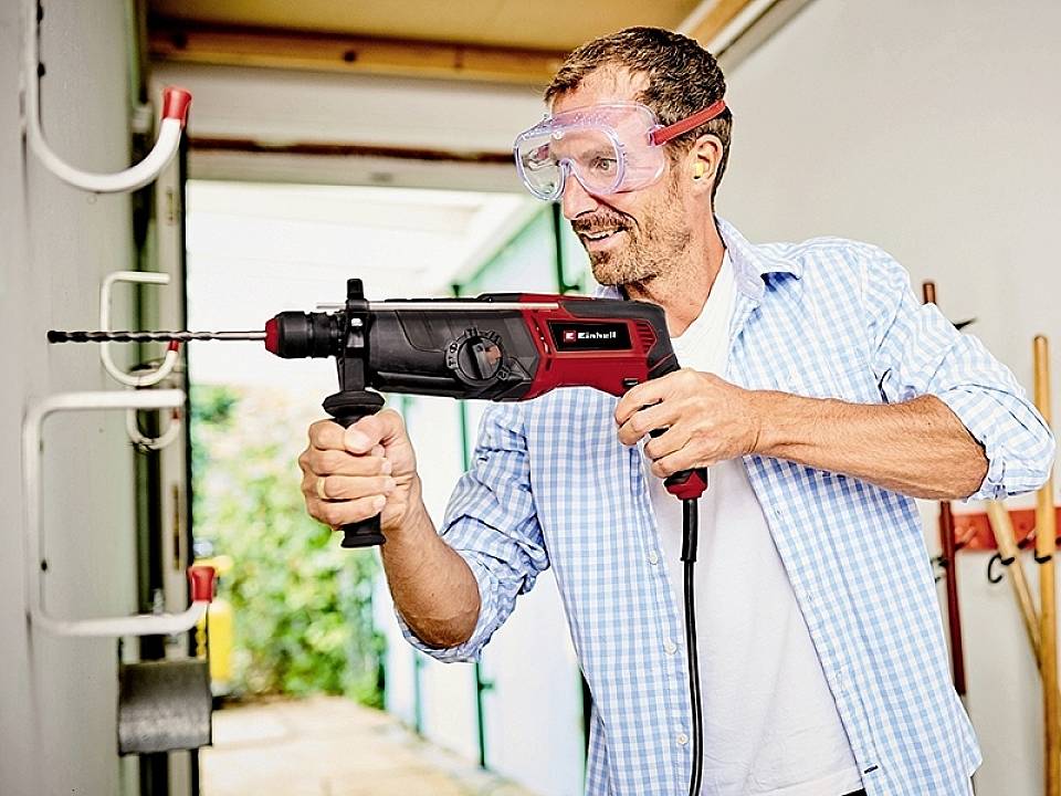 Ein Mann bohrt ein Loch mit einer Bohrmaschine in eine Wand. Er trägt Schutzbrille und lächelt, während er seine Aufgabe ausführt.