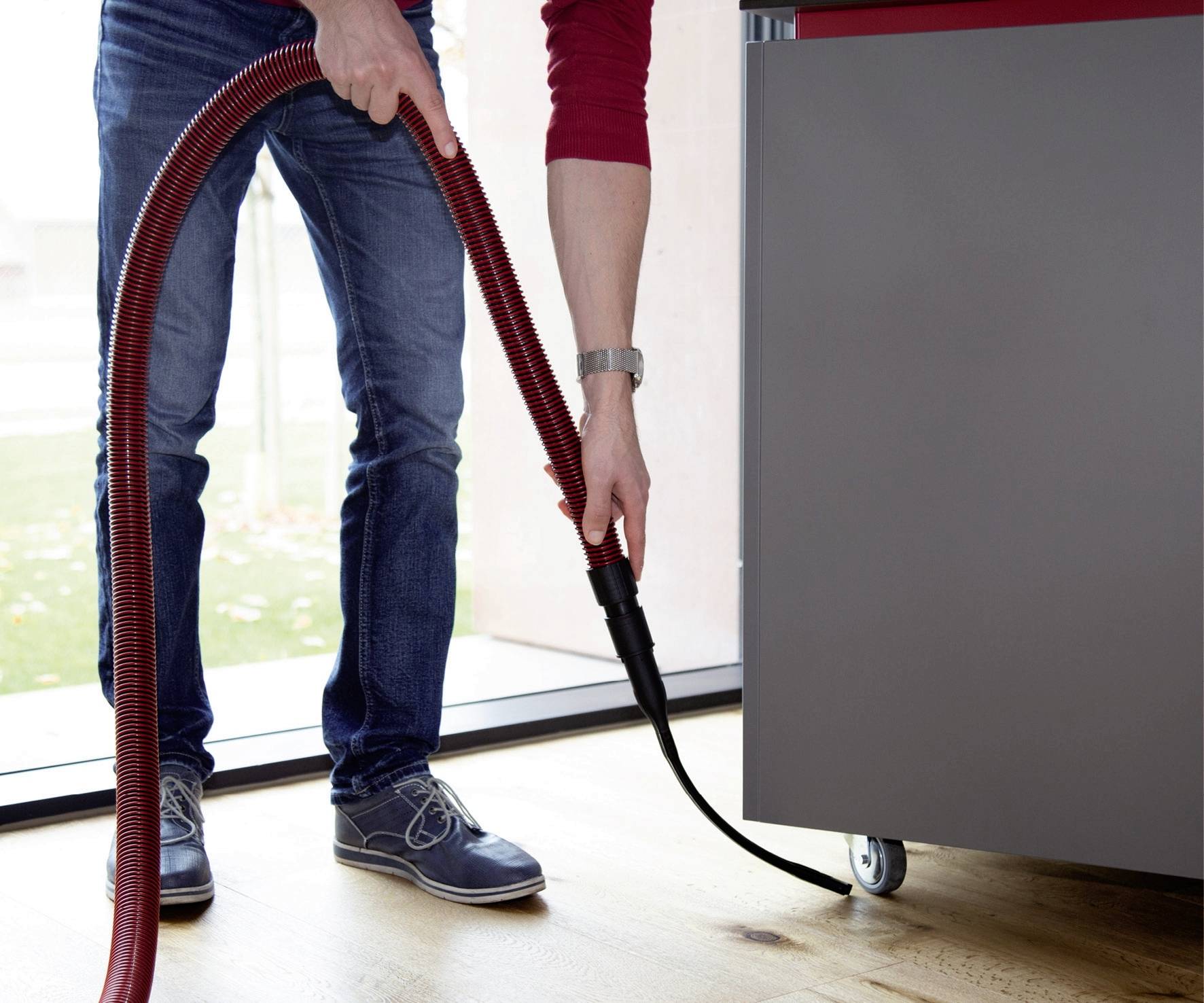 Person in blauer Jeans und roten Pullover saugt mit einem roten Staubsaugerrohr den Boden neben einem grauen Schrank.