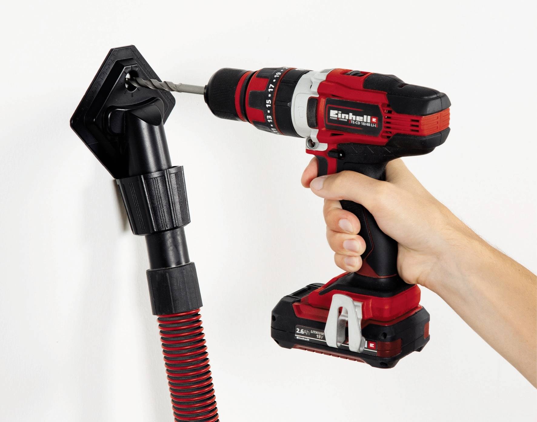 Eine Person bohrt mit einem roten und schwarzen Akkuschrauber ein Loch in eine weiße Wand, während ein Staubsauger den Staub absaugt.