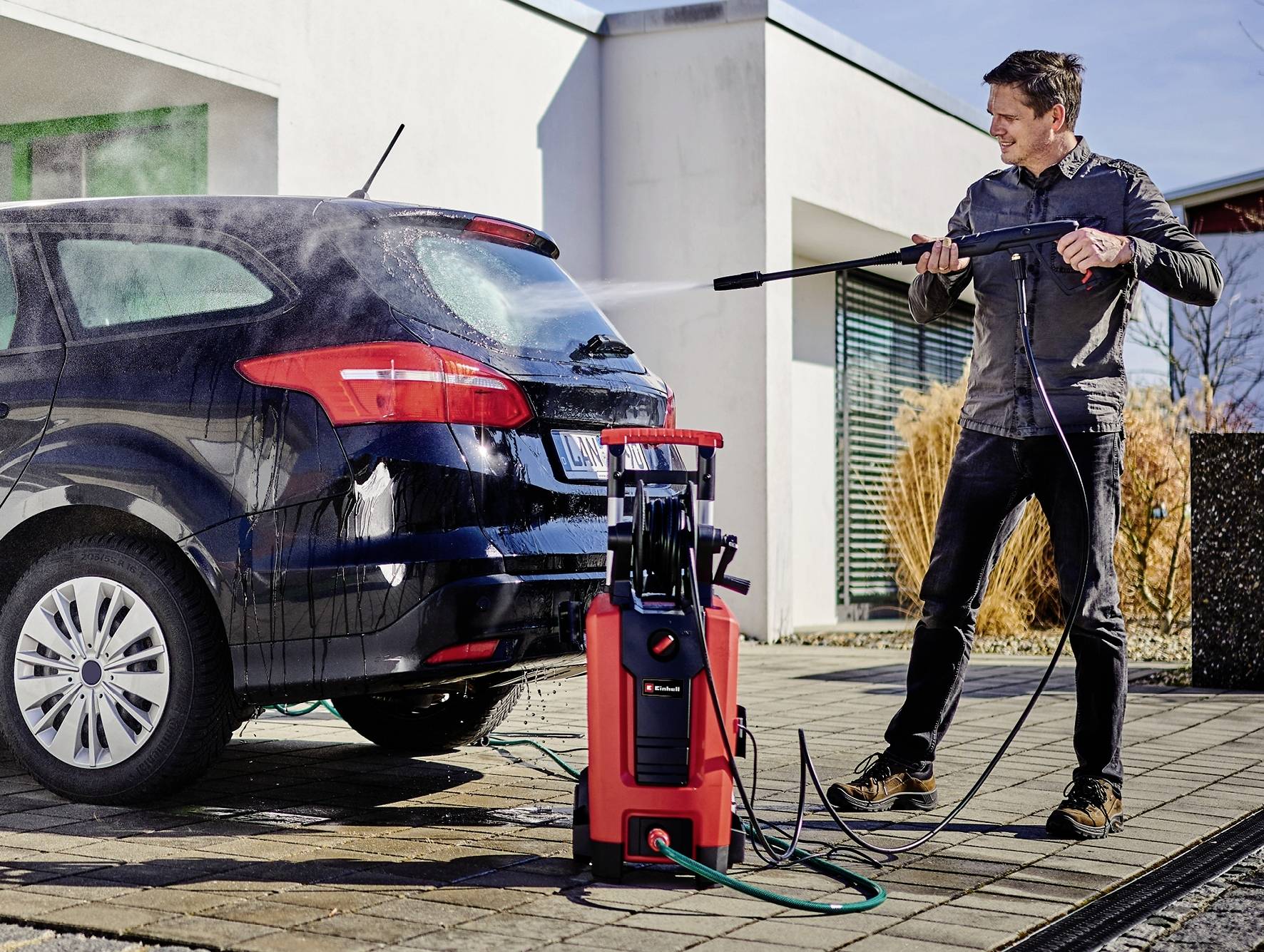 Ein Mann reinigt ein Auto mit einem Hochdruckreiniger auf einer Einfahrt vor einem modernen Gebäude.