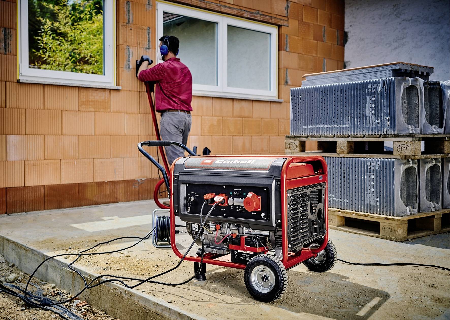 Ein tragbarer Generator steht im Vordergrund einer Baustelle. Im Hintergrund arbeitet eine Person an einer unverputzten Hauswand.