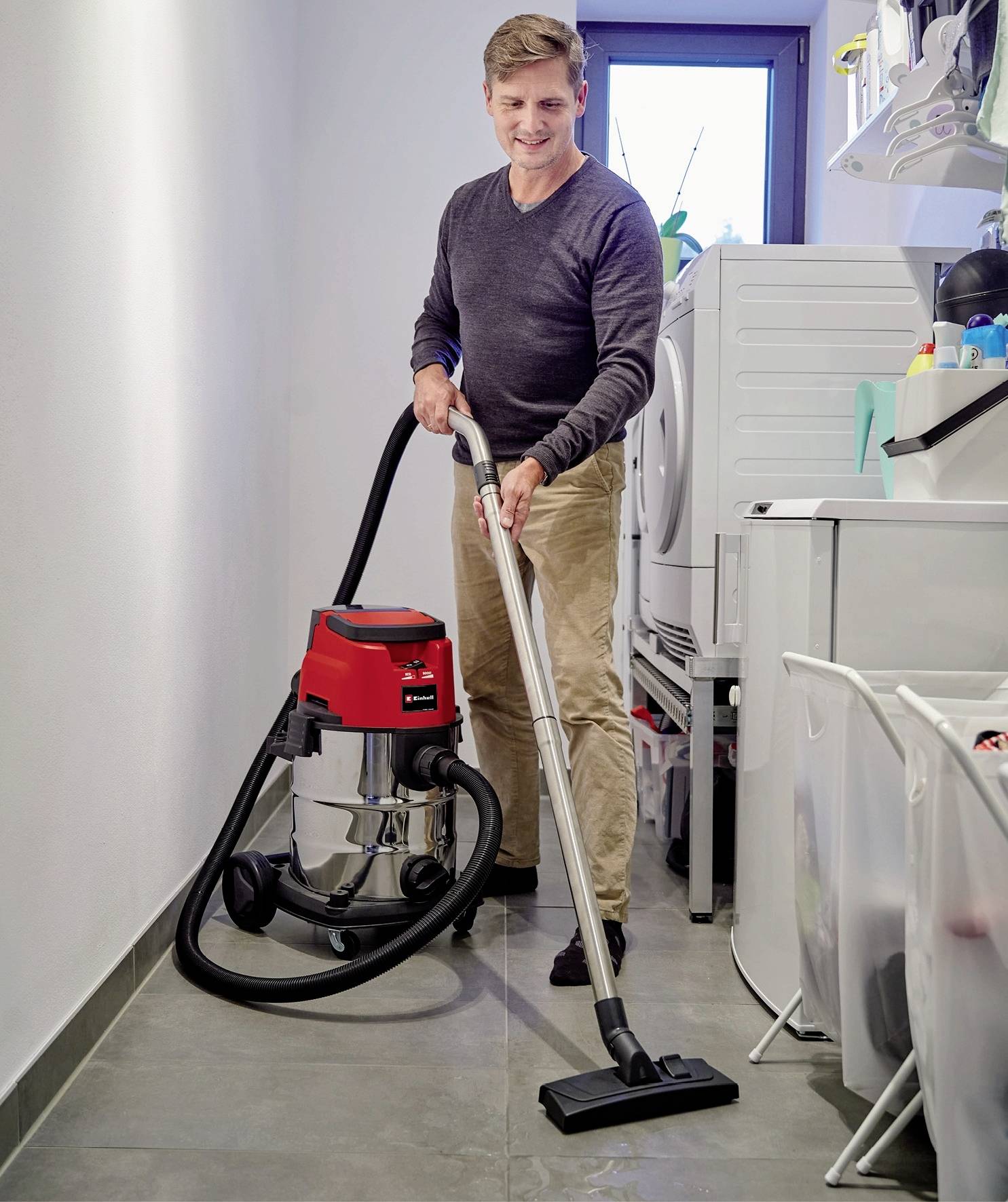 Ein Mann staubsaugt den Boden in einem Hauswirtschaftsraum. Neben ihm stehen eine Waschmaschine und diverse Haushaltsgegenstände.