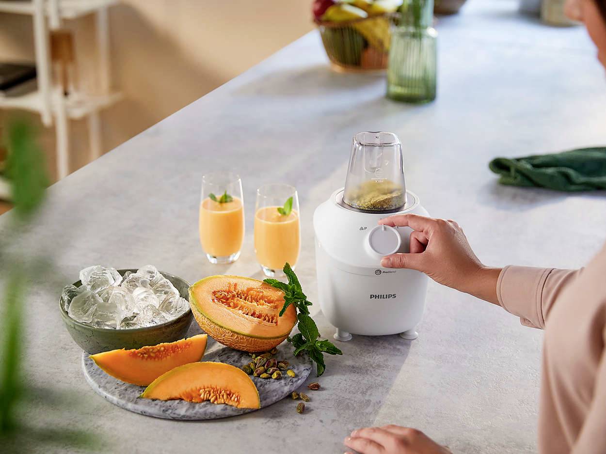 Eine Person bedient einen Küchenmixer auf der Arbeitsfläche. Daneben Melonenscheiben und zwei Gläser mit einem orangen Getränk.