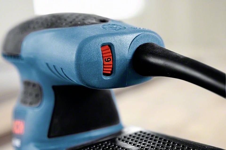 Ein blauer Elektroschleifer mit schwarzem Griff und rotem Geschwindigkeitsregler in der Einstellung 6, liegt auf einem Holztisch.