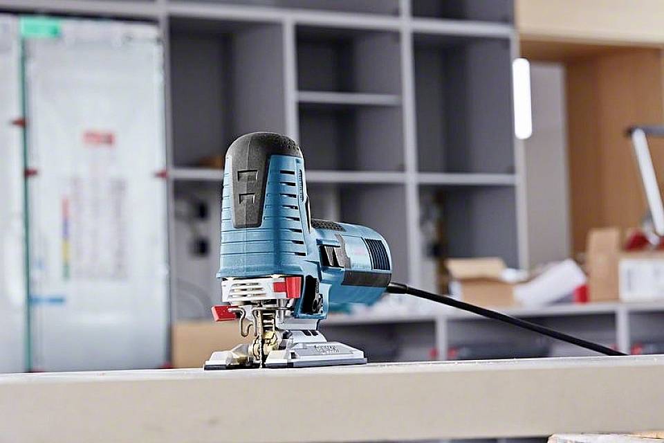 Eine blaue Stichsäge liegt auf einer Arbeitsfläche in einer Werkstatt. Im Hintergrund sind Regale und Werkzeuge sichtbar.
