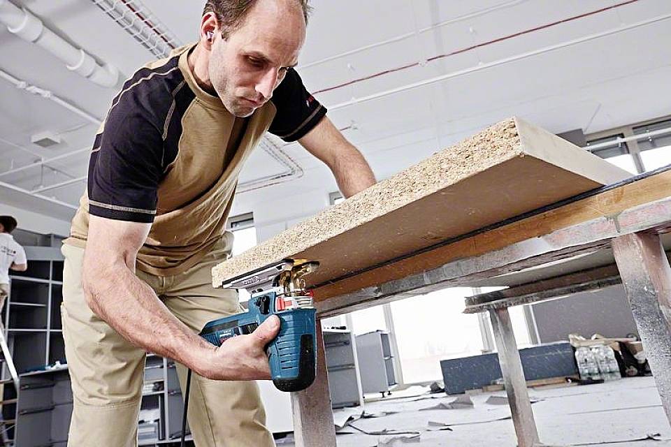 Ein Mann benutzt eine elektrischen Stichsäge, um eine Holzplatte in einem Werkstattbereich zu schneiden. Im Hintergrund sind weitere Werkzeuge sichtbar.