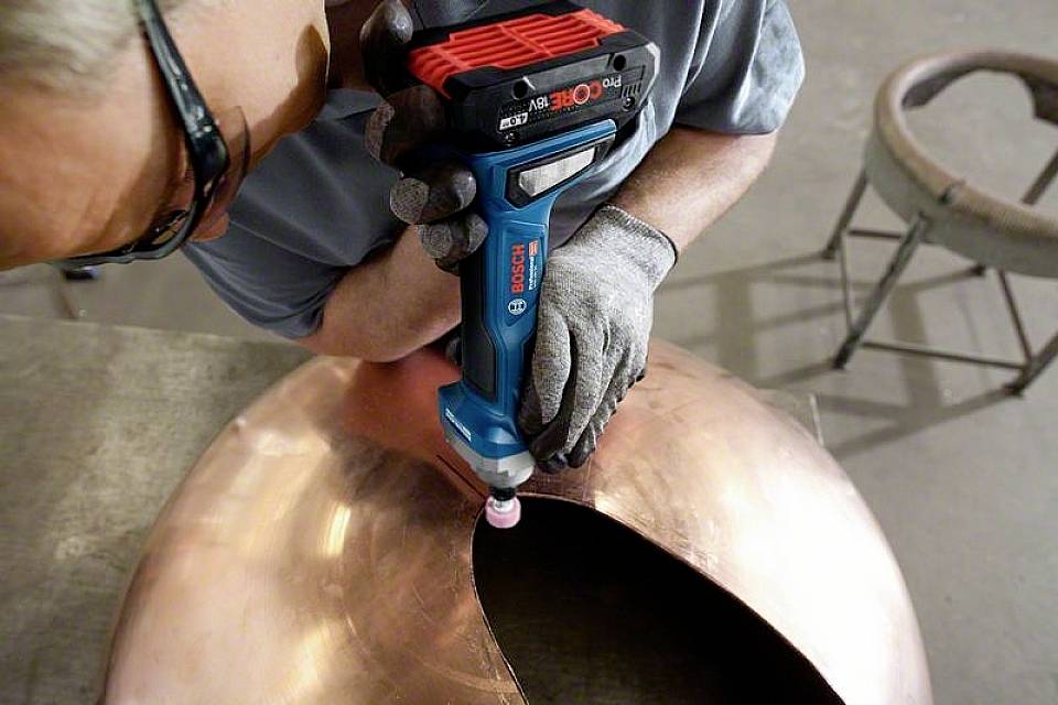 Person schleift eine Kupferkugel mit einem Elektroschleifer in einer Werkstatt. Schutzbrille und Handschuhe werden getragen.