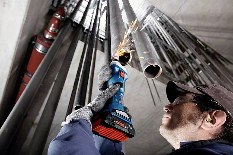 Eine Person arbeitet mit einem elektrischen Werkzeug in einem Industriebereich, Funken fliegen von Metallrohren. Sicherheitsausrüstung ist sichtbar.