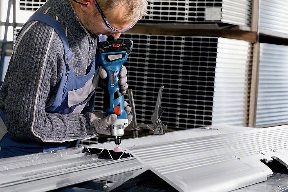 Eine Person bearbeitet ein großes Metallblech mit einem Elektrowerkzeug in einer Werkstattumgebung. Sie trägt Schutzbrille und Arbeitskleidung.