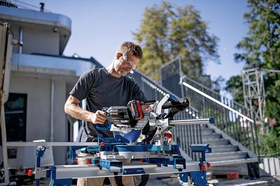 Ein Mann arbeitet im Freien mit einer Kapp- und Gehrungssäge. Er trägt Schutzbrille und Gehörschutz. Im Hintergrund eine Treppe.