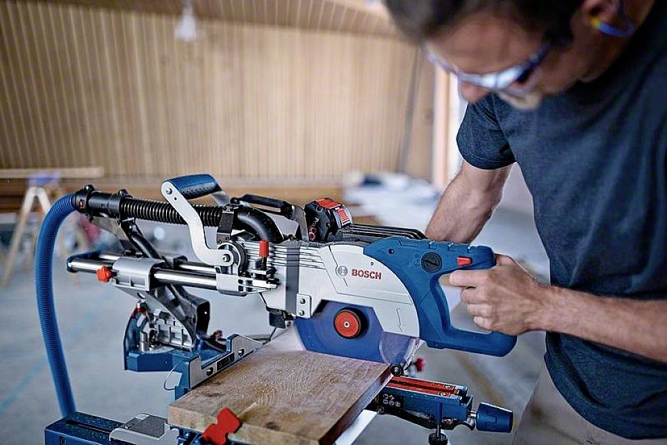 Ein Mann in Werkstatt benutzt eine elektrische Kappsäge, um ein Holzbrett zu schneiden. Die Umgebung wirkt handwerklich und technisch.
