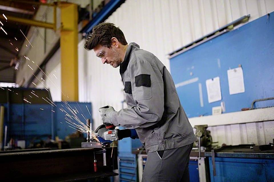Ein Mann in grauer Arbeitskleidung schleift Metall in einer Werkstatt. Funken fliegen umher. Im Hintergrund sind Werkzeuge und blaue Wände zu sehen.