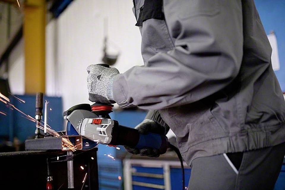 Eine Person in Schutzkleidung schleift Metall mit einem Winkelschleifer, Funken fliegen. Arbeitet in einer Werkstattumgebung.