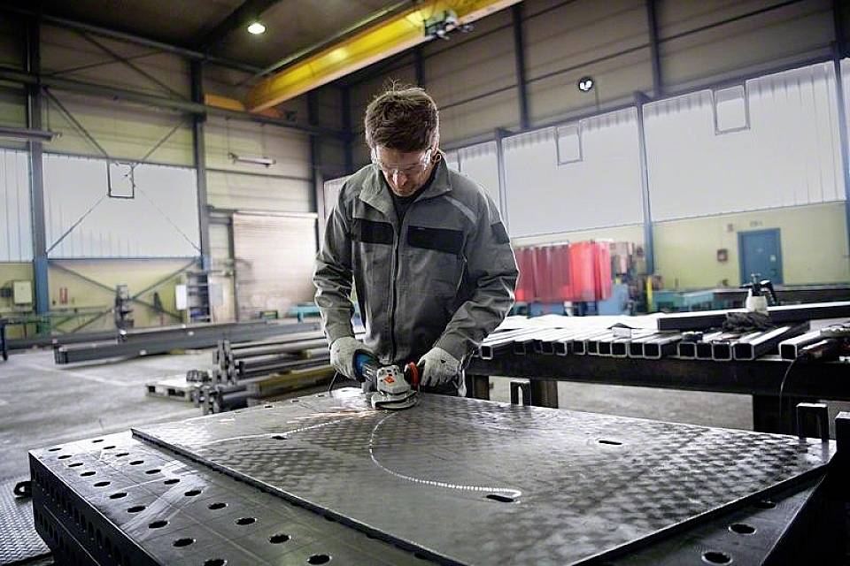 Eine Person schleift eine große Metallplatte in einer Fabrikhalle. Sie tragen Schutzkleidung und arbeiten konzentriert an einem Tisch.