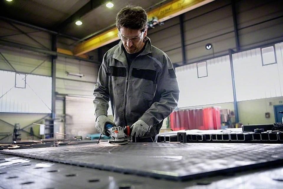 Ein Arbeiter in einer Werkstatt schleift eine Metallplatte mit einem Elektrowerkzeug. Funken fliegen, die Umgebung ist industriell.