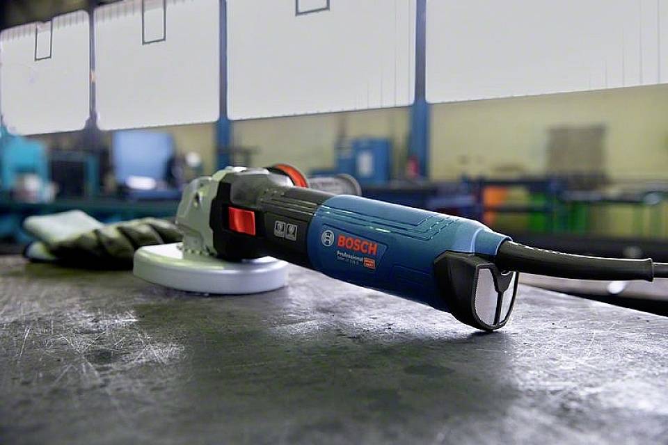 Ein blauer Winkelschleifer von Bosch liegt auf einem Tisch in einer Werkstatt. Im Hintergrund sind unscharfe Maschinen erkennbar.