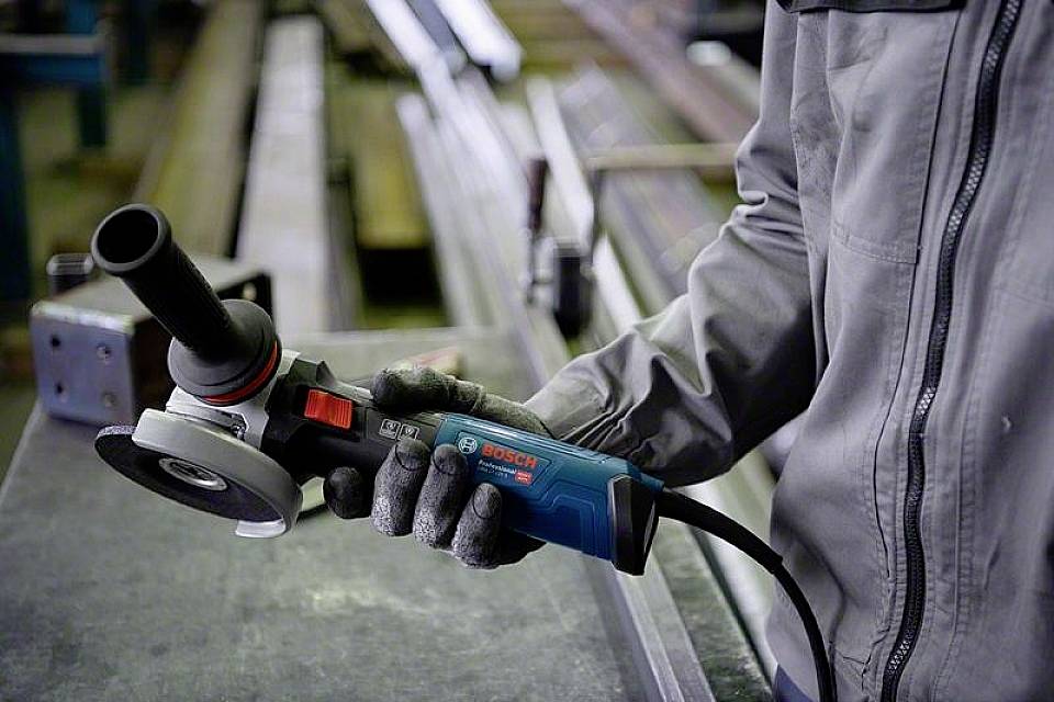 Eine Person hält einen blauen Bosch-Winkelschleifer in einer Werkstatt. Sie trägt Arbeitshandschuhe und graue Schutzkleidung.
