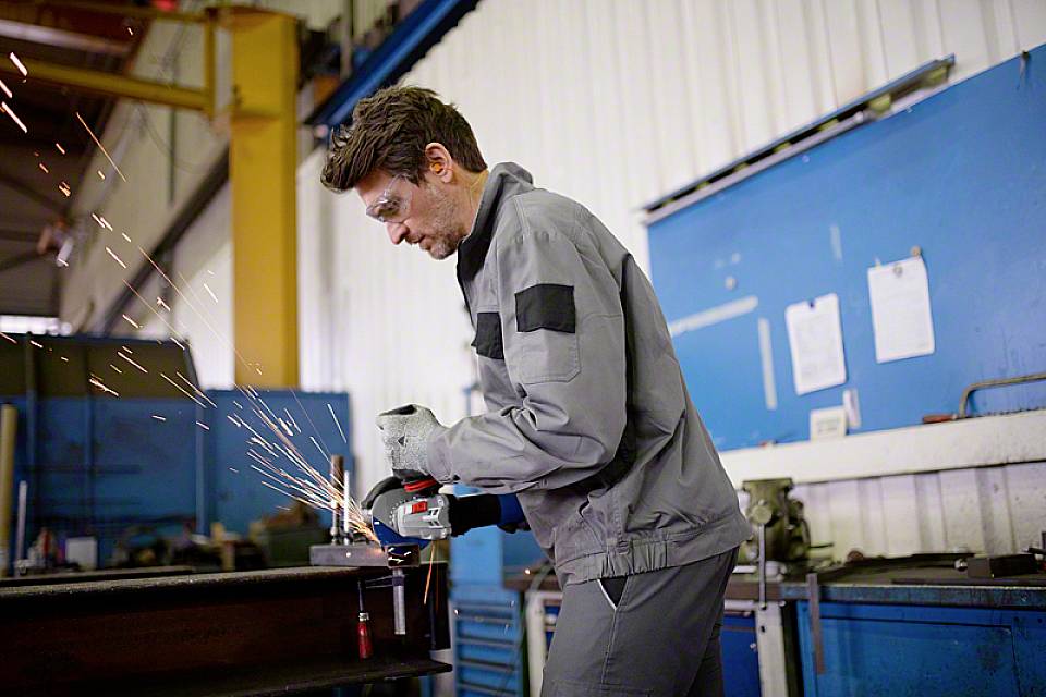 Ein Mann in Schutzkleidung schleift Metall in einer Werkstatt. Funken fliegen von der Schleifmaschine. Hintergrund zeigt Werkstattgeräte.