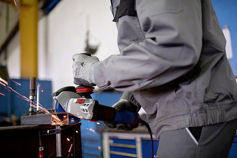 Eine Person in Arbeitskleidung schleift Metall mit einem Winkelschleifer, Funken fliegen. Die Szene zeigt handwerkliche Arbeit in einer Werkstatt.