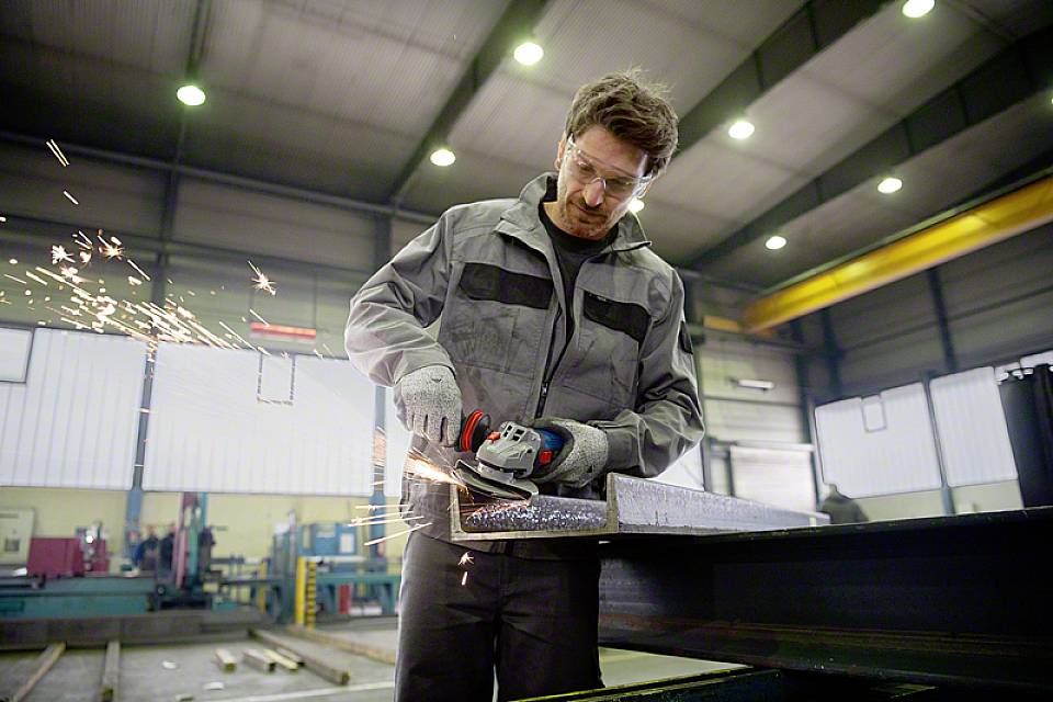 Ein Mann bearbeitet ein Metallstück mit einem Winkelschleifer in einer Werkhalle. Funken fliegen, während er konzentriert arbeitet.