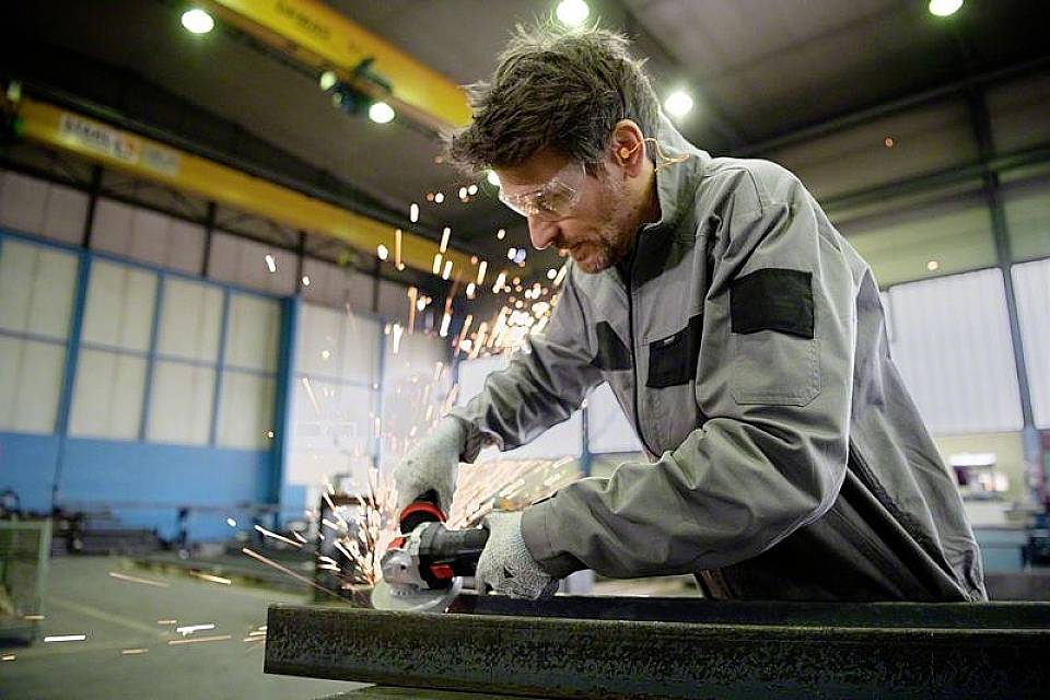 Ein Mann schleift in einer Werkhalle ein Metallstück mit einem Winkelschleifer. Funken sprühen. Er trägt Schutzbrille und Arbeitskleidung.