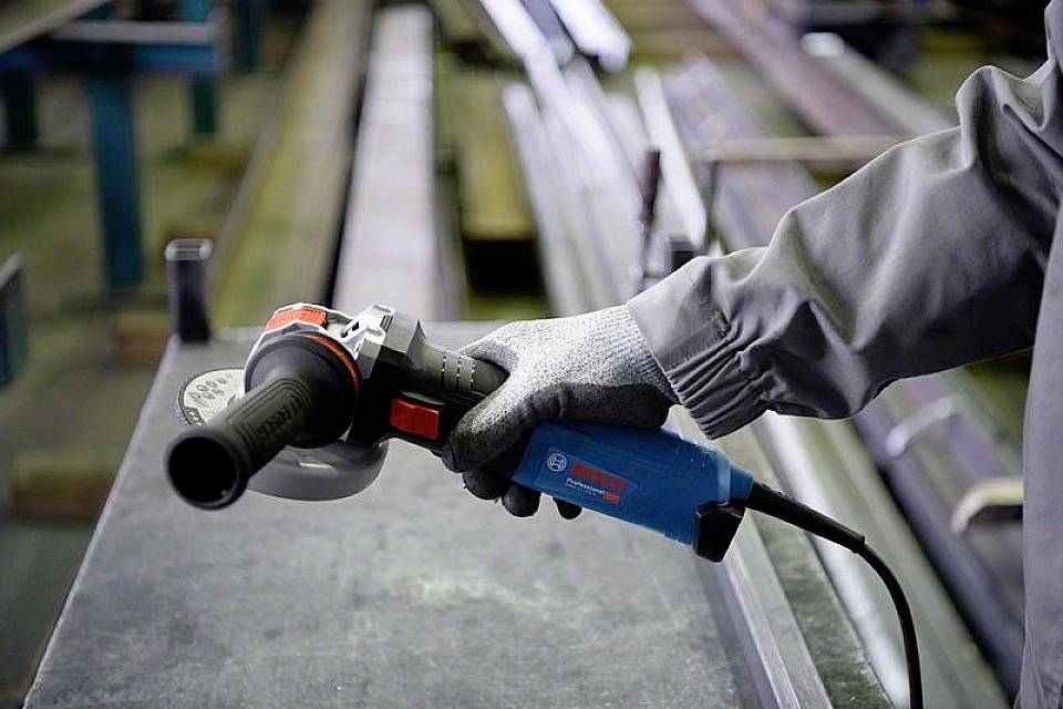 Eine Person in Arbeitskleidung hält einen Winkelschleifer über einem Metalltisch. Schutzhandschuhe sind zu sehen.