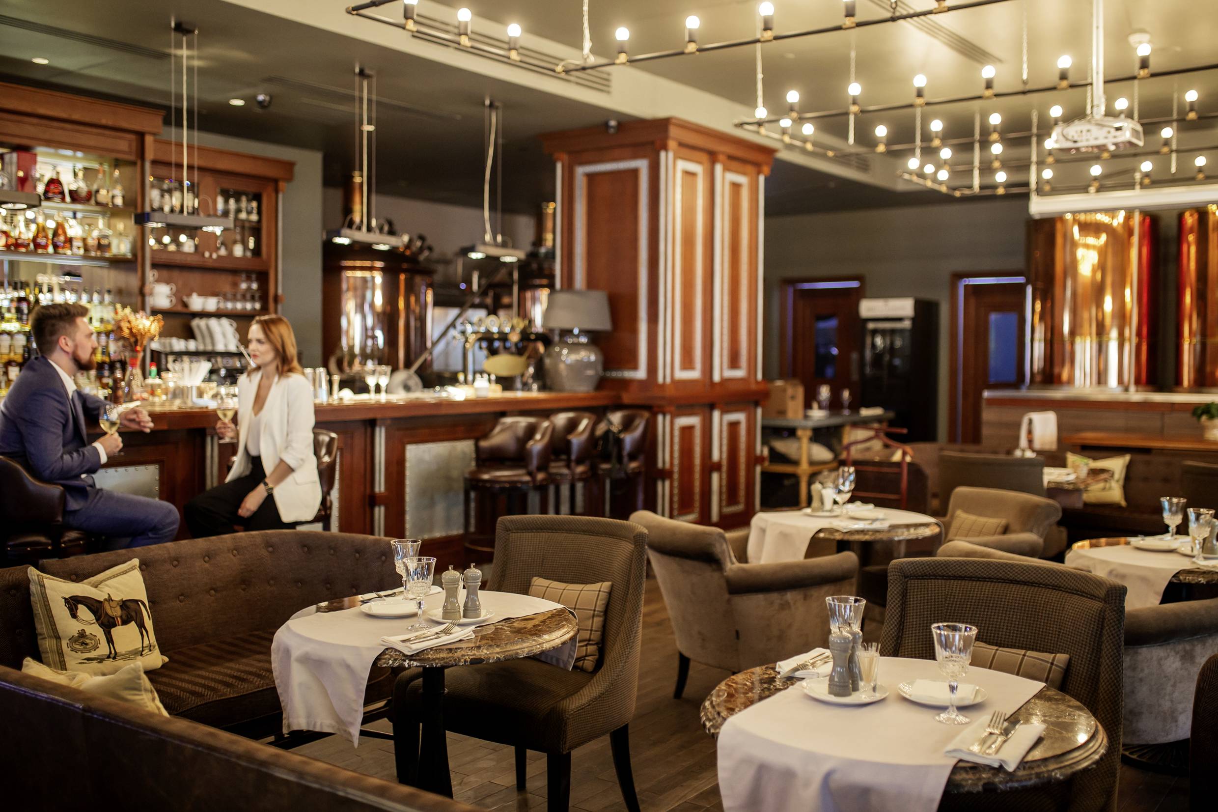 Eine stilvolle Bar mit Holzdekor und modernen Lampen. Vorne befinden sich gemütliche Tische, im Hintergrund unterhält sich ein Paar an der Theke.