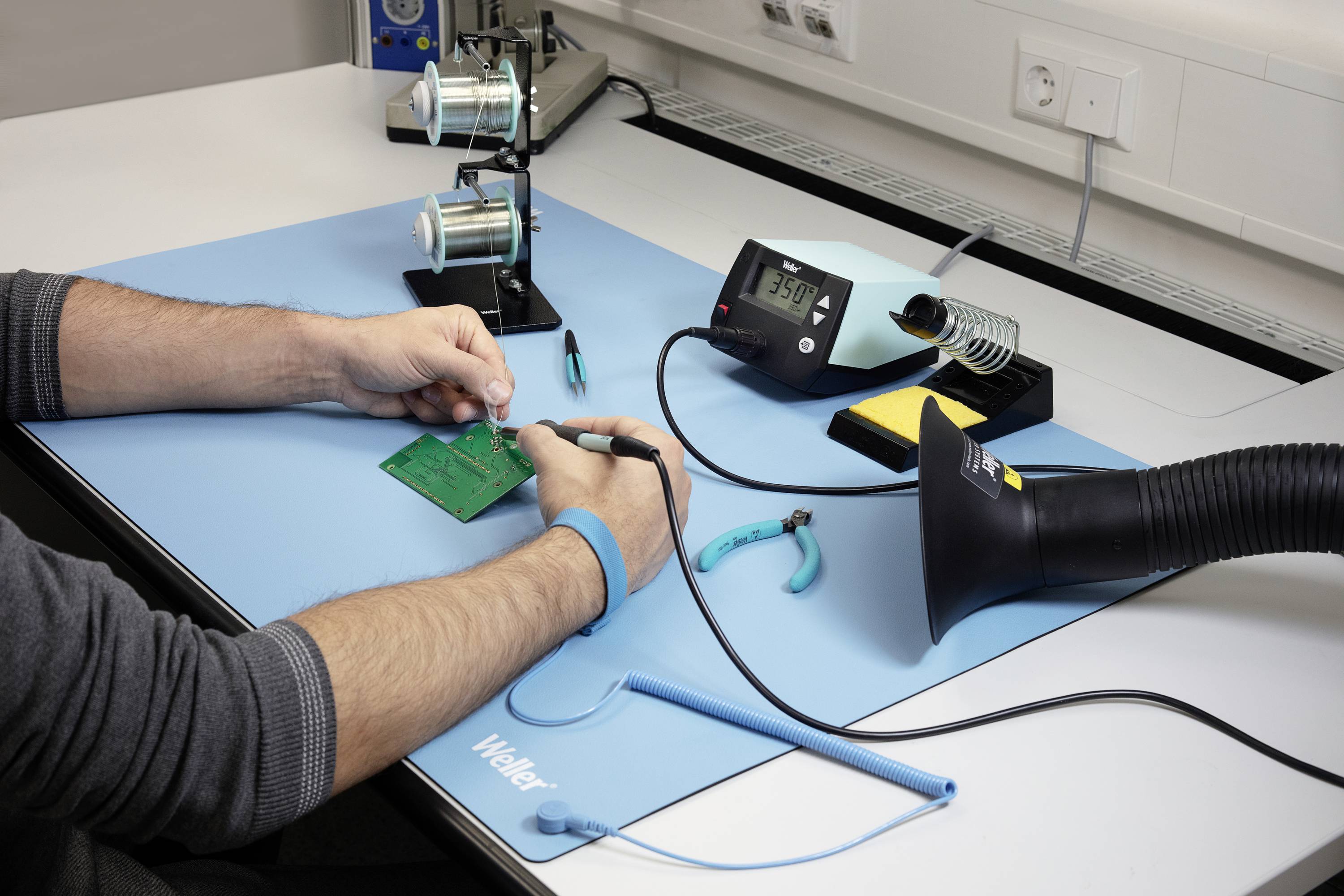 Person arbeitet an einer Leiterplatte mit Lötkolben auf Werkbank. Es gibt einen Lüfter zur Dampfabsaugung und ein Multimeter zur Spannungsprüfung.