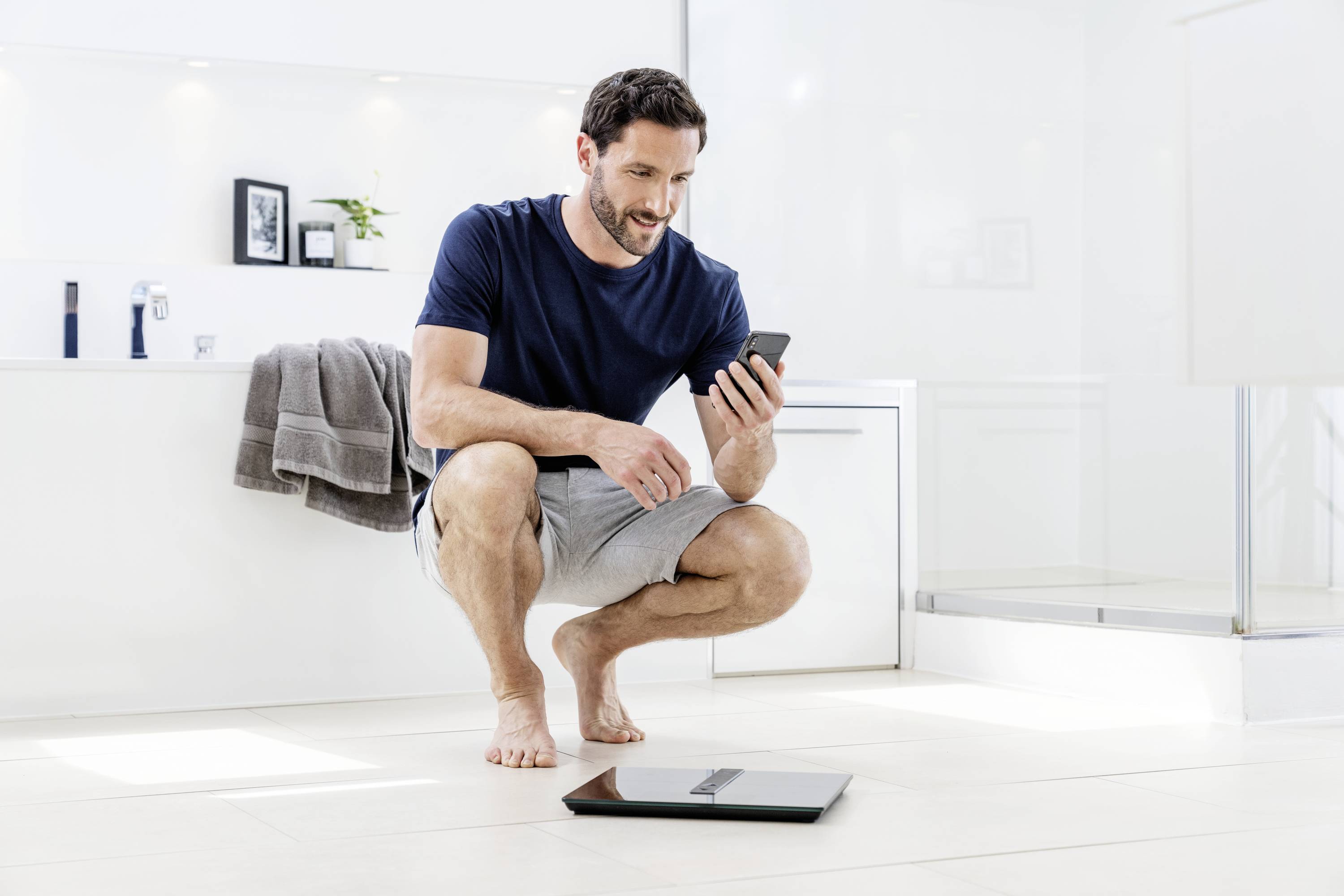 Ein Mann sitzt in einem Badezimmer und betrachtet sein Smartphone. Vor ihm auf dem Boden liegt eine digitale Waage.