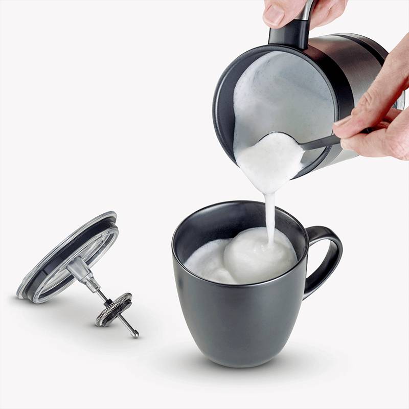 Eine Person gießt aufgeschäumte Milch aus einem Kännchen in eine schwarze Tasse. Daneben liegt ein Deckel mit Filter.