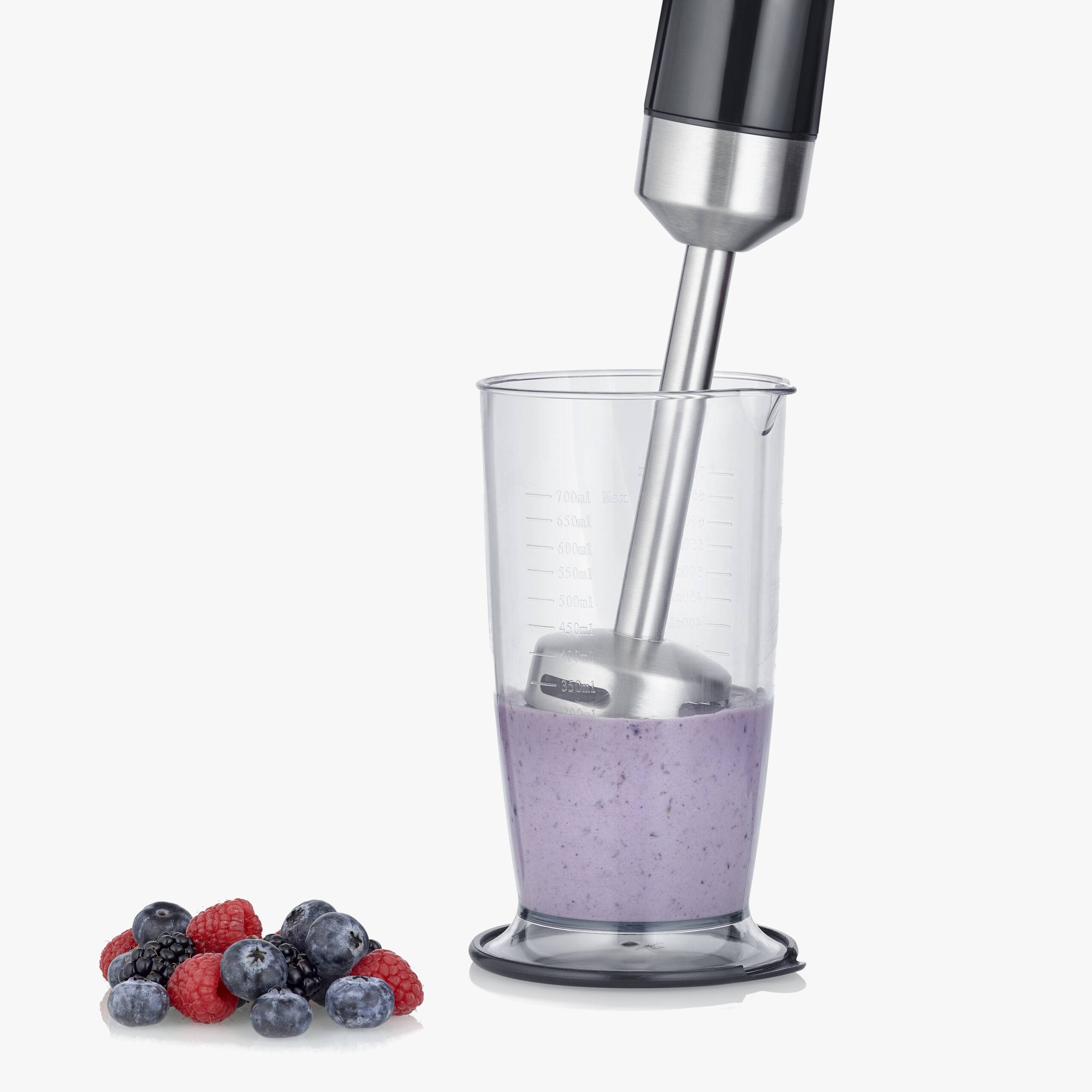 Ein Stabmixer püriert einen lila Smoothie in einem Messbecher. Daneben liegen einige Himbeeren und Blaubeeren auf einem Tisch.