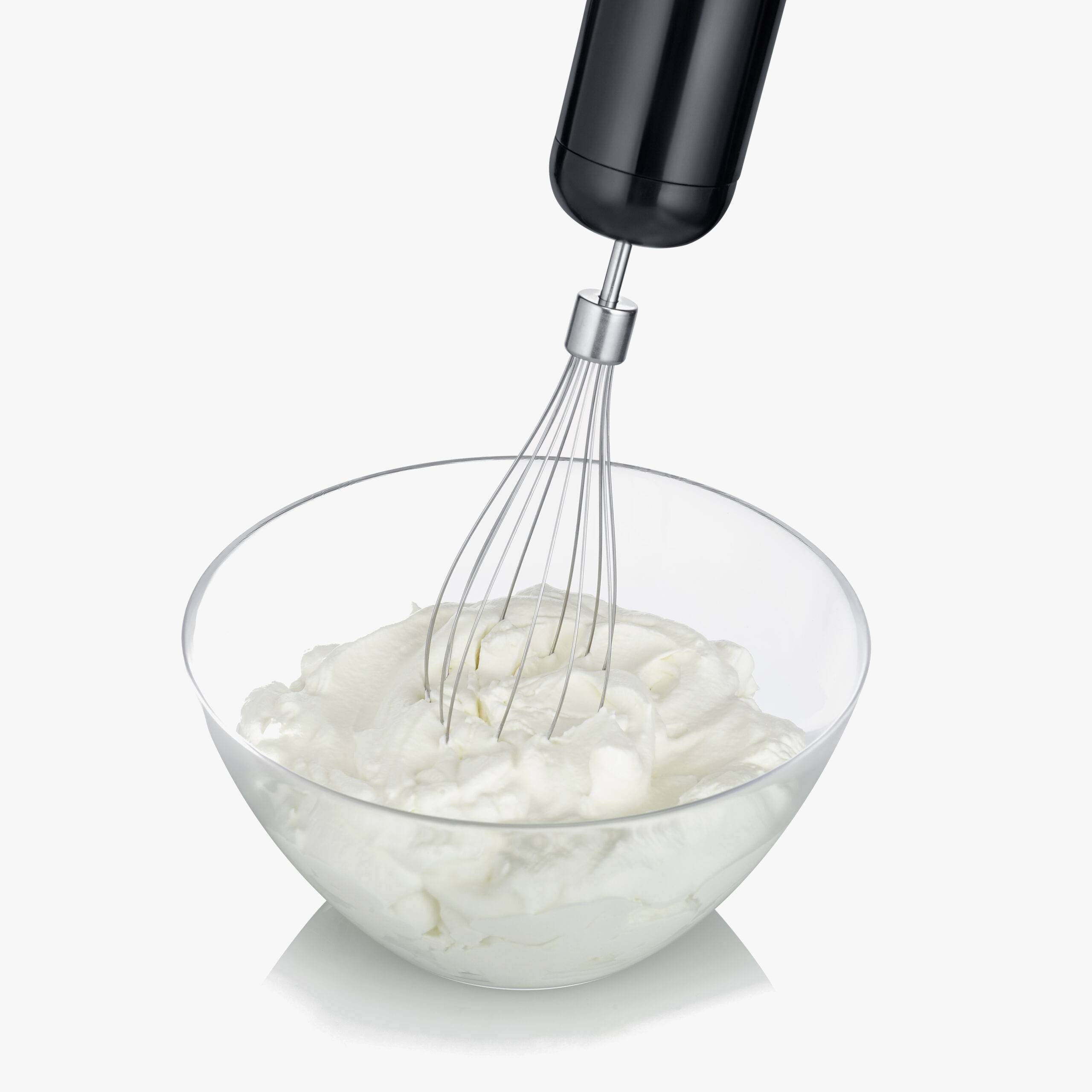 Ein elektrischer Handmixer schlägt Sahne in einer transparenten Schüssel. Die Sahne beginnt fest zu werden.