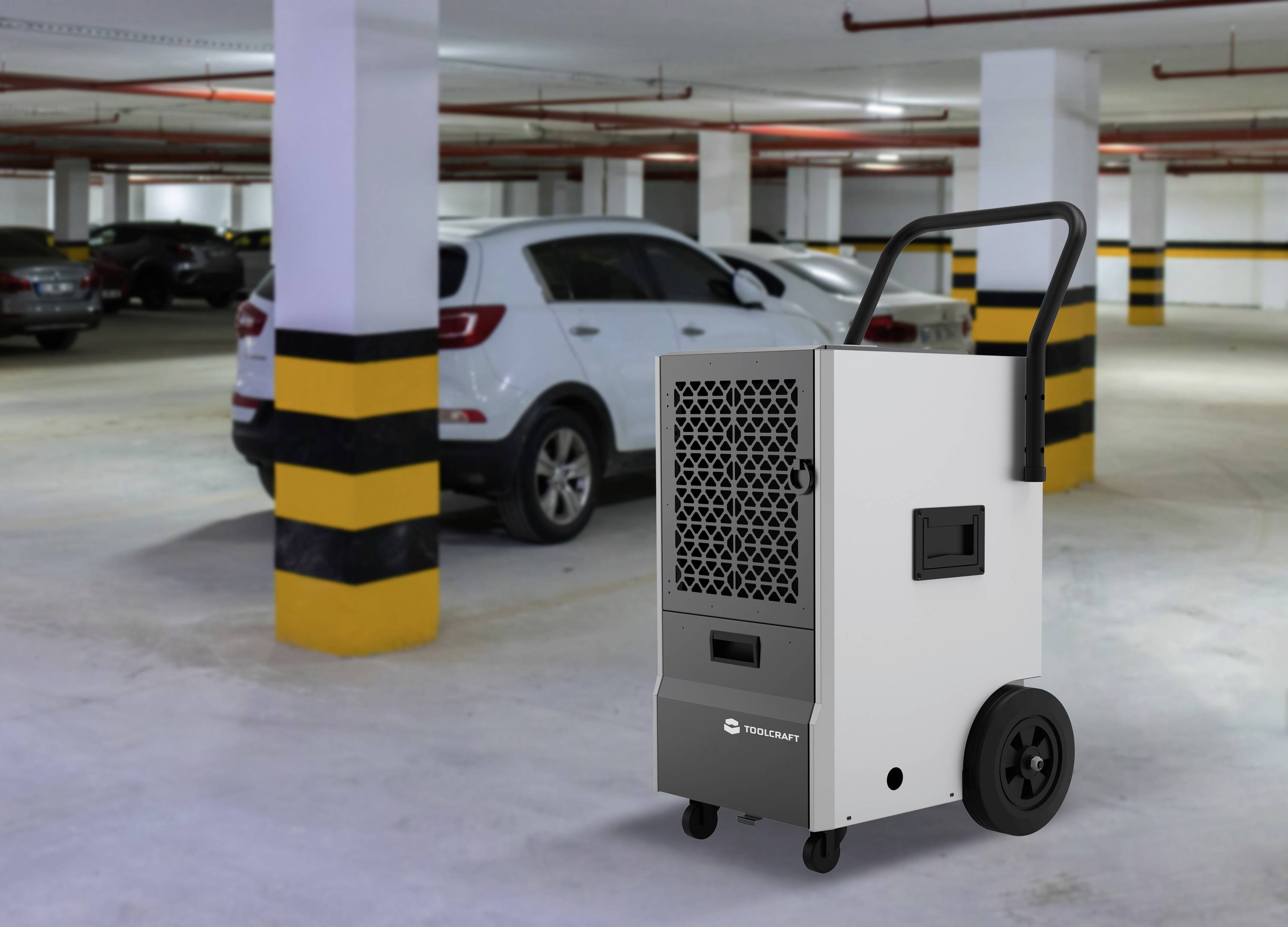 Ein weißer Luftentfeuchter steht in einem leeren Parkhaus neben einem geparkten Auto. Der Hintergrund zeigt weiße und gelbe Wände.