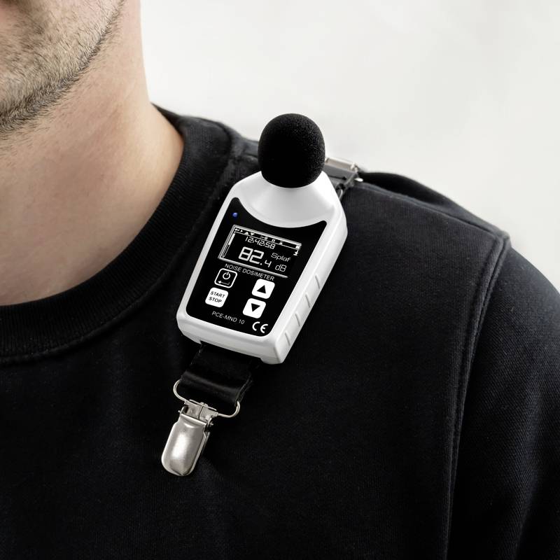 Ein Mann trägt ein Dezibel-Messgerät an seinem Pullover. Das Display zeigt 82,4 dB mit einem Batteriestatus und Mikrofon-Icon an.