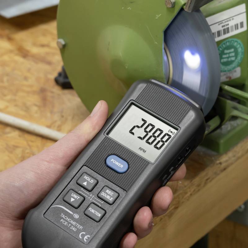 Eine Hand misst die Drehzahl einer rotierenden Schleifscheibe mit einem Tachometer. Das Gerät zeigt '2988 RPM' an.