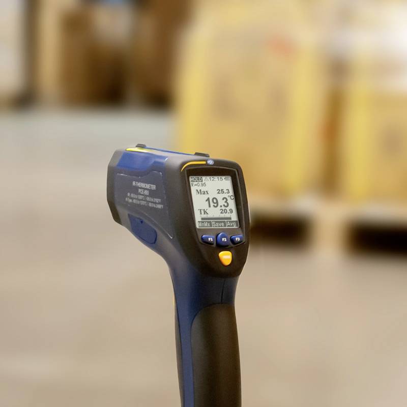 Ein Infrarotthermometer zeigt auf ein Objekt. Der Bildschirm zeigt eine maximale Temperatur von 25,5°C. Im Hintergrund unscharfe Kartons.
